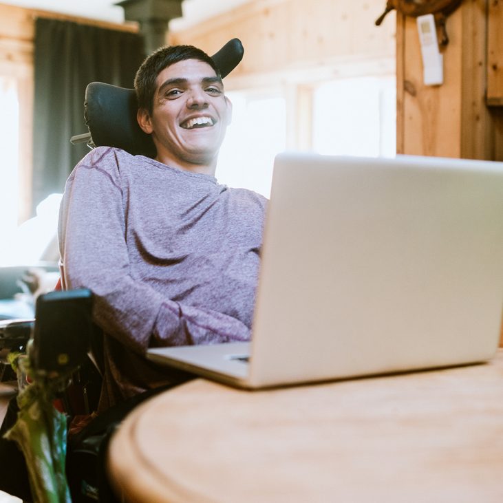 Junger Mann mit Behinderung sitzt lächelnd am Tisch mit Laptop